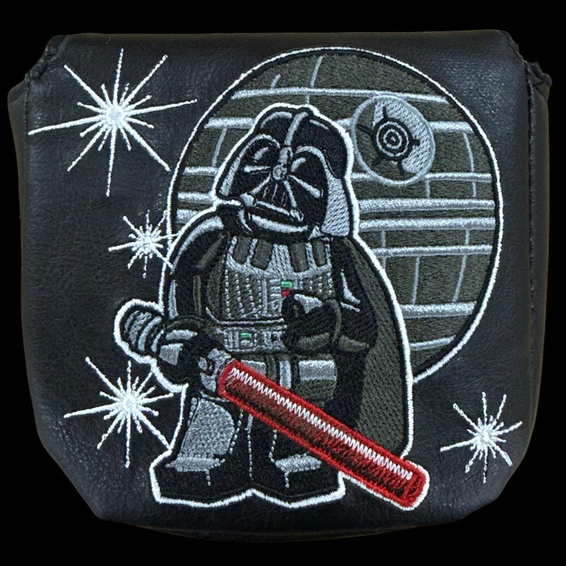 May include: A black leather golf putter headcover featuring an embroidered design of a stylized character in a black helmet and suit holding a red lightsaber, set against a circular space station background with white stitched starburst accents.