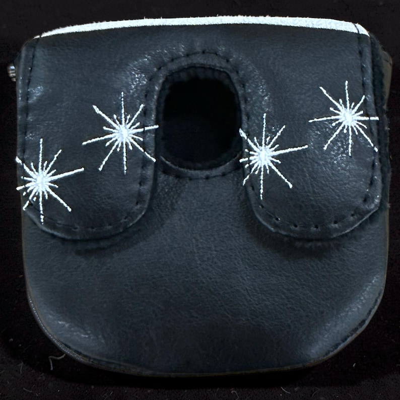 May include: A black leather putter cover featuring a white embroidered starburst pattern across the front flap. The cover has a central cutout opening and a smooth, matte finish, providing a protective layer for a golf club head.