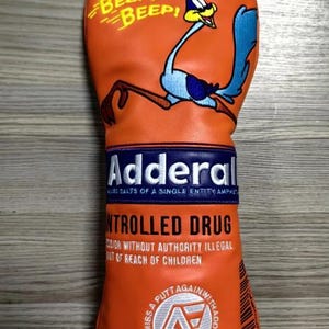 Può includere: Copertura arancione per la testa della mazza da golf con un personaggio dei cartoni animati di Road Runner e la scritta "BEEP! BEEP!". La copertura presenta anche la scritta "Adderall" e "CONTROLLED DRUG".
