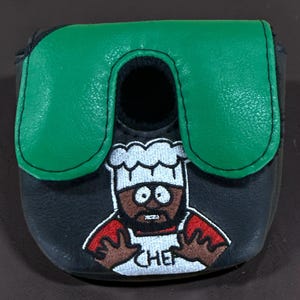 May include: A black mallet putter headcover featuring a green top panel and an embroidered graphic of a cartoon chef wearing a white hat and apron with the word CHEF written on it. The design includes red sleeves and brown skin tones.