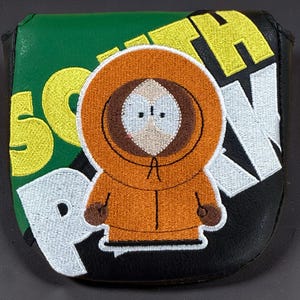 Könnte beinhalten: Ein schwarz-grüner Golf-Putter-Schlägerhaube mit einem gestickten Charakter in einem orangefarbenen Kapuzenparka. Der Schriftzug SOUTH PARK ist in großen gelben und weißen Buchstaben auf den Hintergrund gestickt und umrahmt die zentrale Figur.