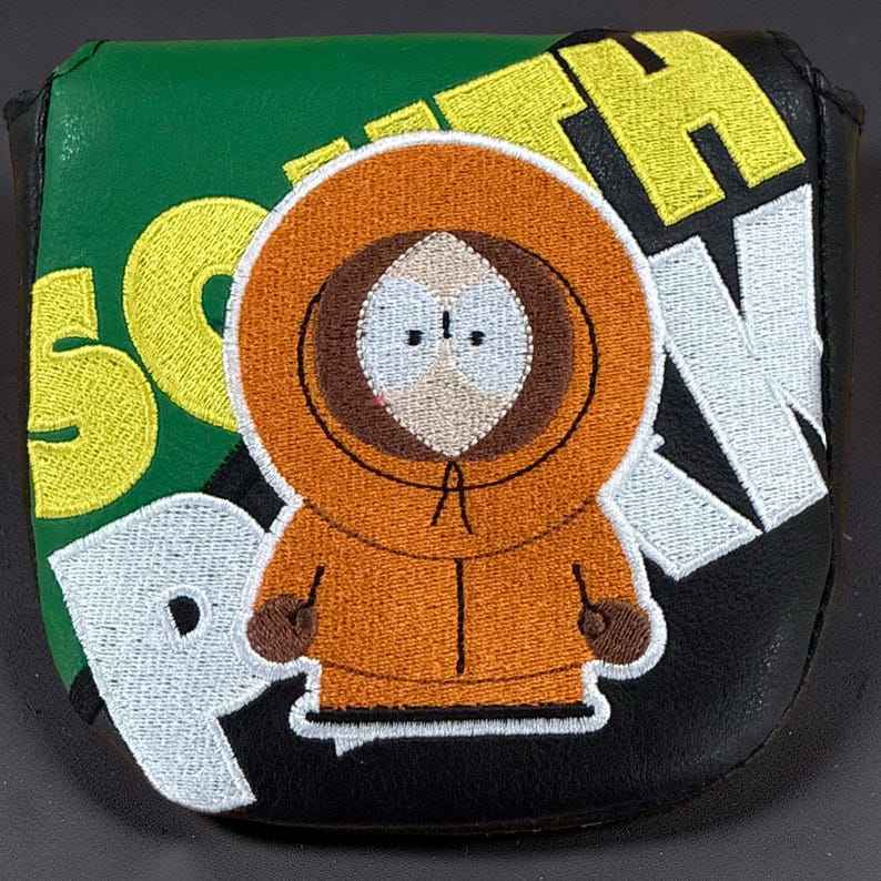May include: A black and green golf putter headcover featuring an embroidered character wearing an orange hooded parka. The text SOUTH PARK is stitched in large yellow and white letters across the background, surrounding the central character figure.