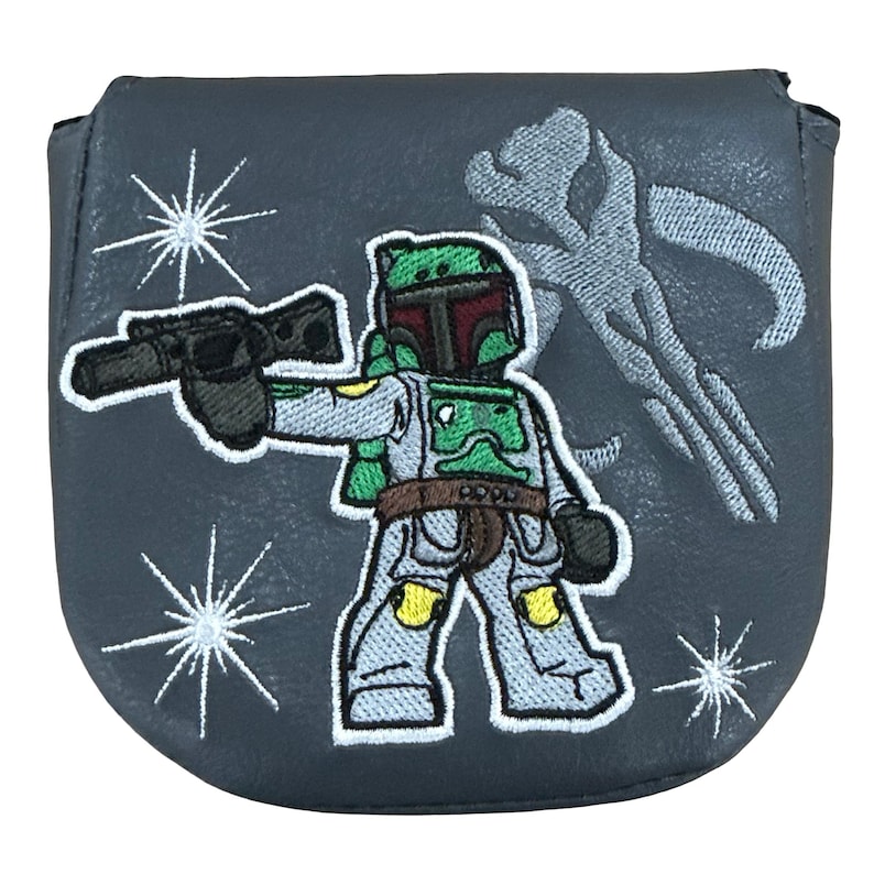 K&ouml;nnte beinhalten: Ein dunkelgrauer Putter-Schl&auml;gerhaube mit einer gestickten Grafik einer stilisierten gepanzerten Figur, die einen Blaster h&auml;lt. Das Design umfasst ein gro&szlig;es Totenkopf-Emblem im Hintergrund und drei wei&szlig;e Sternmuster auf der glatten grauen Oberfl&auml;che.