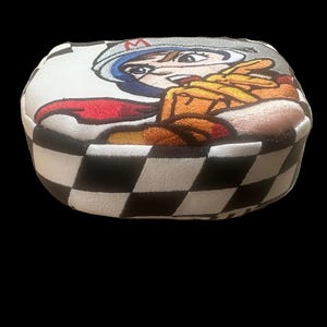 Puede incluir: Una funda para la cabeza del palo de golf con un dise&ntilde;o de corredor de dibujos animados. La funda presenta un patr&oacute;n de cuadros blancos y negros en los laterales y una imagen bordada colorida de un corredor en la parte superior. El corredor lleva casco y traje de carreras.