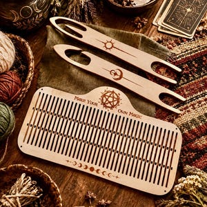 Pode incluir: Um pente de tecelagem de madeira clara e duas lançadeiras. O pente apresenta ranhuras verticais, um design de fases lunares e o texto "MAKE YOUR OWN MAGIC" com um pentagrama. As lançadeiras estão gravadas com um sol e uma lua crescente.