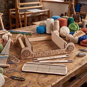 May include: A wooden weaving loom kit featuring a frame with floral and animal engravings, two long shuttles, and a rectangular heddle. The set is surrounded by various spools and balls of yarn in blue, green, orange, and beige on a wooden table.