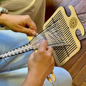 May include: A wooden weaving tool with a sunburst design is used to create a woven textile. The textile features black, white, and lavender threads. The weaver's hands are visible, manipulating the threads and tool.