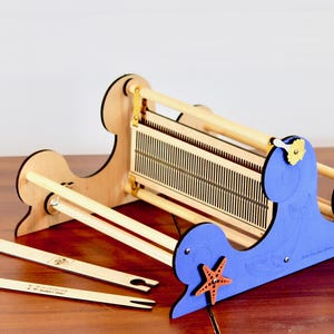 May include: A wooden loom with a blue and white sea-themed design. The loom has a wooden frame and a blue base with a starfish and other sea creatures. The loom is made of wood and has a blue and white paint finish. The loom is designed for weaving and has a wooden shuttle and a wooden heddle.
