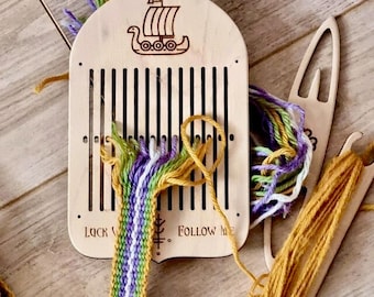 May include: A wooden weaving loom with a ship illustration and the text "LUCK FOLLOW ME". The loom has vertical slots and is partially threaded with colorful yarn in shades of purple, green, and yellow. A weaving shuttle is also visible.