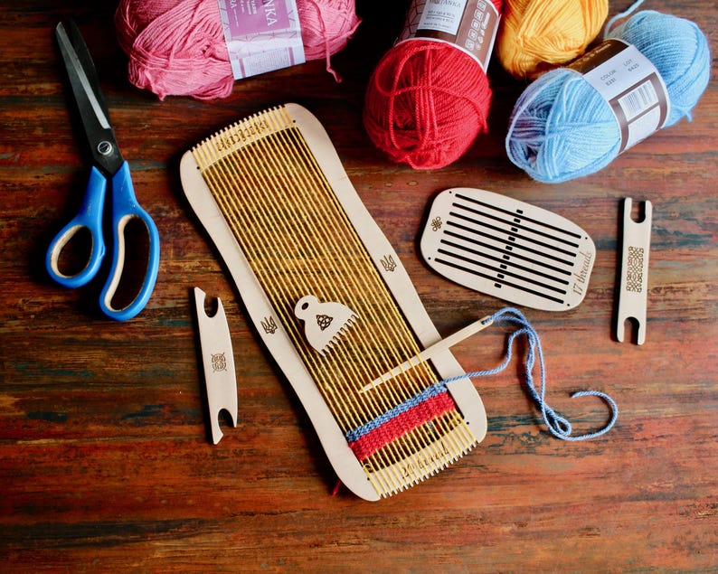 May include: A wooden loom with a blue, red, and white woven piece on it. There are four balls of yarn in the background: pink, red, yellow, and blue. There are also two wooden weaving tools and a pair of blue scissors.