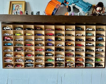 Puede incluir: Una estantería de madera con 72 compartimentos individuales, cada uno con un coche de juguete en miniatura. Los coches son de varios colores, incluyendo rojo, azul, verde, amarillo y negro. La estantería está montada en una pared.