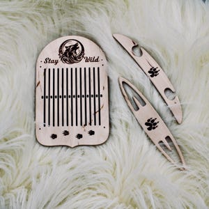 May include: Wooden loom with a wolf design and the text "Stay Wild." The loom is made of light wood and has a natural finish. It comes with two additional wooden tools.