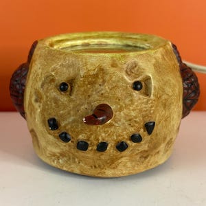 May include: A tan, textured wax warmer shaped like a snowman head with a carrot-colored nose, black dot eyes, and a wide, black-dotted smile. The top features a gold-toned rim, and dark brown, textured ear muffs are visible on the sides.