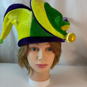 May include: A colorful jester hat with a tri-color design of green, yellow, and purple. The hat has a velvet texture and features a gold ball at the end of one of the points. The hat is designed for festive occasions.