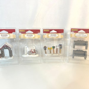 May include: Four miniature Christmas village accessories in clear plastic packaging. The accessories include a red and brown arched entrance, a red bicycle, a clothesline with colorful laundry, and two decorative benches.
