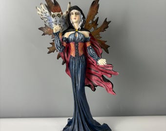 May include: A figurine of a fairy with an owl. The fairy wears a dark blue dress with a red and orange bodice and a red cape. The fairy has brown wings and a black hood. The owl is white, brown, and blue.