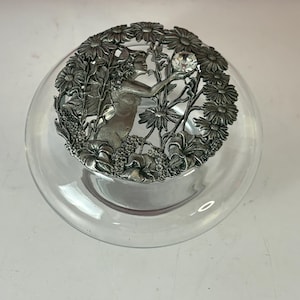 May include: A clear glass bowl with a decorative metal lid. The lid features a detailed relief of a woman reaching for a crystal, surrounded by flowers. The metal has a silver finish.