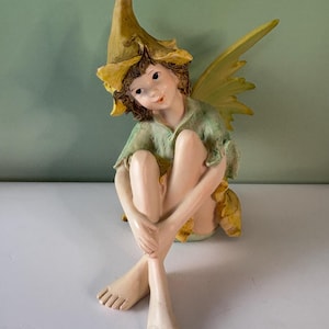 Puede incluir: Una caprichosa figura de hada con un sombrero en forma de flor amarilla y alas a juego. El hada lleva una túnica verde y está sentada con las piernas cruzadas. La figura tiene una expresión serena y está pintada en tonos suaves y naturales.
