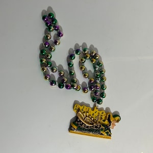 May include: A Mardi Gras necklace with green, gold and purple beads. The necklace has a Mardi Gras themed charm featuring a jester and musical notes.
