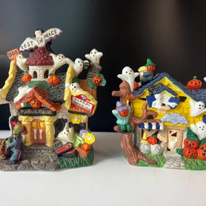 May include: Two Halloween-themed ceramic houses. One is a funeral parlor with ghosts and pumpkins, the other a floral horror shop. Both have detailed designs, including signs, ghosts, and jack-o'-lanterns. The houses are brightly colored.