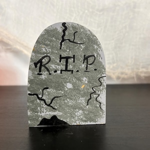 May include: A gray tombstone with the letters "R.I.P." in black. The tombstone has a cracked texture and is decorated with black squiggly lines, creating a spooky Halloween decoration.