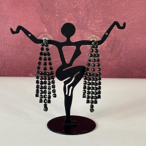 May include: Black rhinestone dangle earrings on a black metal stand. The earrings have multiple strands of small, square-cut rhinestones. The stand is a silhouette of a person in a dancing pose, with a round base.