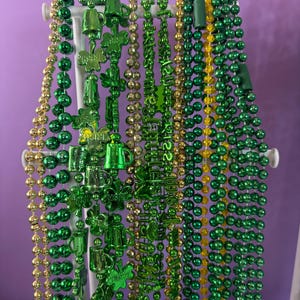 May include: A display of St. Patrick's Day beads in various shades of green and gold. Some beads feature shamrocks, beer mugs, and the text "Irish 22". The beads are hanging on a white display stand against a purple background.