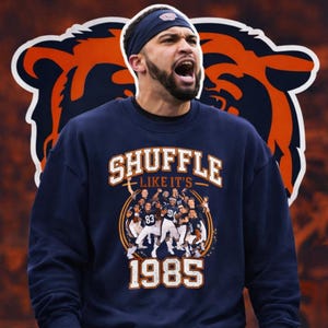 May include: A navy blue crewneck sweatshirt featuring a graphic print of a football team in white uniforms. The text reads SHUFFLE LIKE IT'S 1985 in white and orange lettering. The background shows a large orange and navy blue bear head logo.