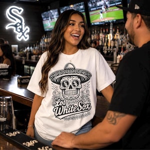 May include: A white short-sleeved t-shirt featuring a black graphic print of a sugar skull wearing a sombrero. The skull has the Chicago White Sox logo on its forehead. Below the skull, the text reads Los White Sox in a stylised script font.