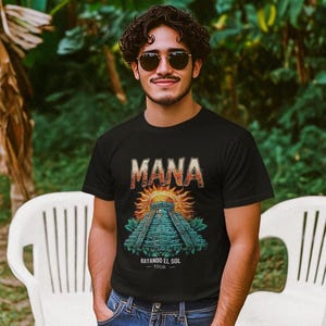 May include: A black t-shirt featuring a graphic of a Mayan pyramid and the word "MANA" in large, white letters. The graphic also includes a sun with rays and the text "Rayando El Sol - Tour".