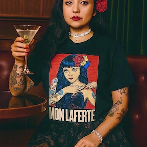 May include: A black short-sleeved t-shirt featuring a graphic print of a person with blue hair and a red flower in their hair. The text MON LAFERTE is printed in bold white capital letters at the bottom of the rectangular graphic design.