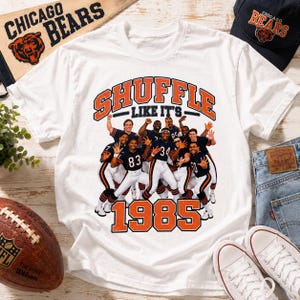 May include: A white t-shirt featuring a graphic of the 1985 Chicago Bears football team. The text reads SHUFFLE LIKE IT'S 1985 in orange and navy blue. The design includes multiple players in uniform, with the team name and year prominently displayed.