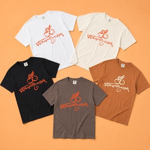 May include: Five short-sleeved crew neck t-shirts in white, cream, black, dark grey, and tan. Each shirt features a central graphic of a stylised flower above two circular shapes and the cursive text Tropi-Gusta in a contrasting colour.
