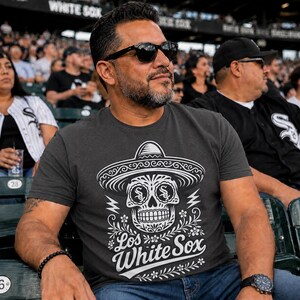 May include: A charcoal grey short-sleeved t-shirt featuring a white graphic of a sugar skull wearing a sombrero. The skull has Chicago White Sox logos in the eye sockets. Below the skull, the text reads Los White Sox in a stylised script font.