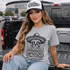 May include: A heather grey t-shirt featuring a black graphic of a sugar skull wearing a sombrero with the text Los White Sox. The shirt is paired with a white and black trucker hat that also displays a sugar skull graphic and the text Los White Sox.