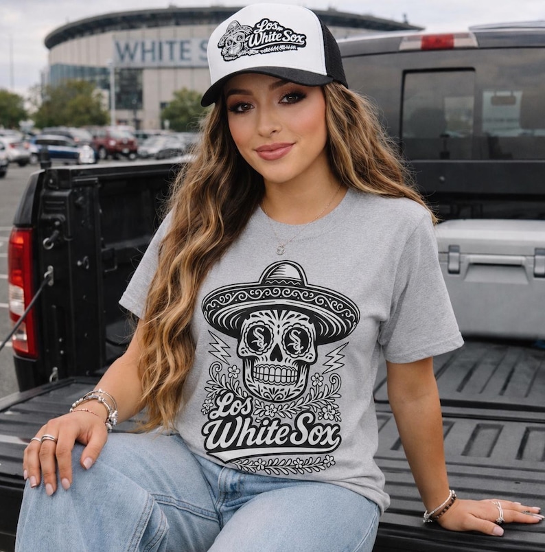 May include: A heather grey t-shirt featuring a black graphic of a sugar skull wearing a sombrero with the text Los White Sox. The shirt is paired with a white and black trucker hat that also displays a sugar skull graphic and the text Los White Sox.
