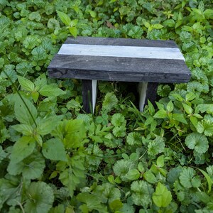 Puede incluir: Un pequeño taburete rectangular de madera con un acabado gris oscuro y una franja blanca horizontal pintada en el centro de la superficie superior. El taburete cuenta con cuatro patas cortas y una parte superior plana, colocado horizontalmente.
