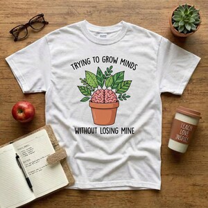 May include: A white t-shirt featuring a graphic of a terracotta pot containing a human brain with green leaves sprouting from the top. The text above the graphic reads TRYING TO GROW MINDS and the text below reads WITHOUT LOSING MINE.