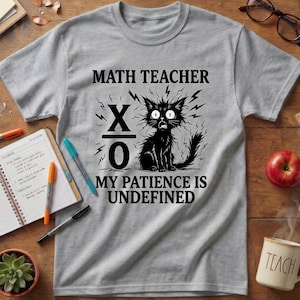 May include: A heather gray short-sleeve t-shirt featuring a black graphic of a wide-eyed cat with jagged lines. The text reads MATH TEACHER, X over O, and MY PATIENCE IS UNDEFINED. The design is centered on the chest of the crew neck garment.