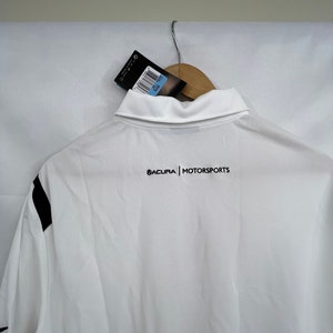 Puede incluir: Polo blanco con detalles negros en las mangas. La camisa tiene cuello y el texto "ACURA | MOTORSPORTS" impreso en negro en la espalda. Una etiqueta cuelga del cuello.