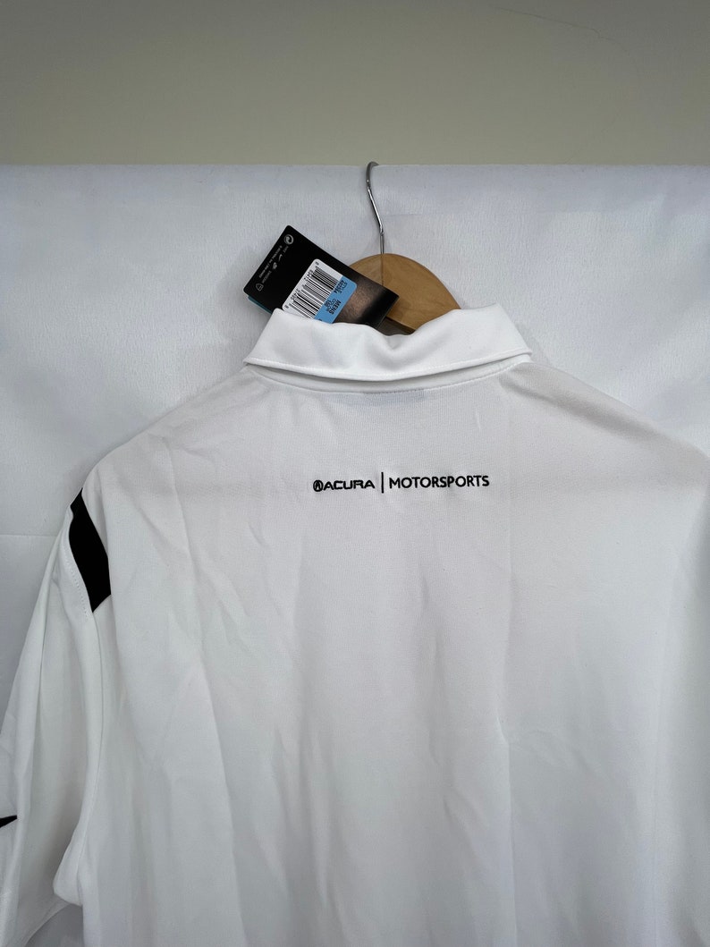 Puede incluir: Polo blanco con detalles negros en las mangas. La camisa tiene cuello y el texto "ACURA | MOTORSPORTS" impreso en negro en la espalda. Una etiqueta cuelga del cuello.