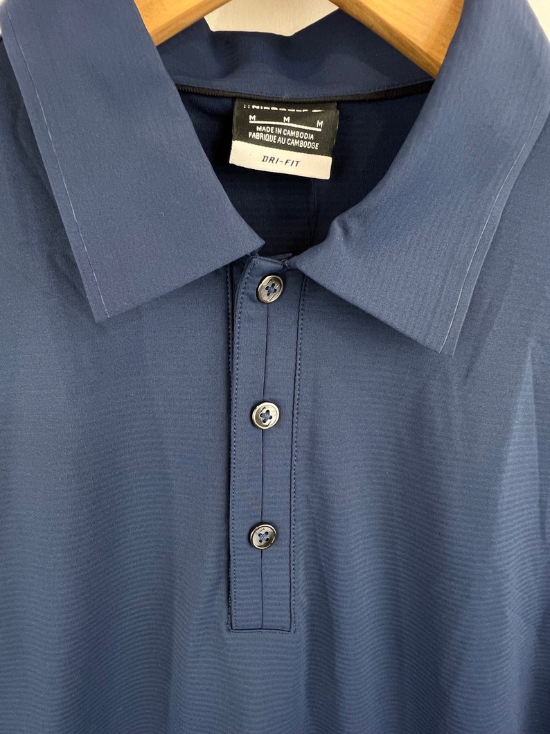 May include: A blue collared shirt with a button-up closure. The shirt has a Dri-FIT label and says "Made in Cambodia, Fabriqu&eacute; au Cambodge" on the tag. The shirt has a size M label.