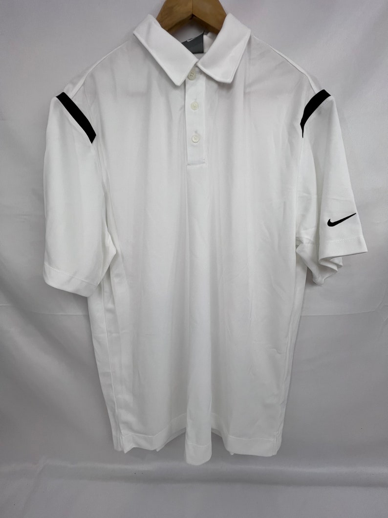 Puede incluir: Polo blanco con cuello y mangas cortas. Presenta detalles negros en los hombros y un logotipo de Nike negro en la manga. La camisa tiene una tapeta de tres botones.