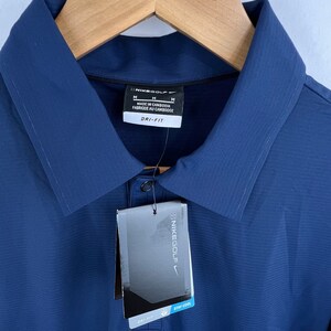 May include: A navy blue golf shirt with a white tag that says "Nikegolf" and "Dri-Fit Stay Cool". The shirt has a button-up collar and a small pocket on the left chest.