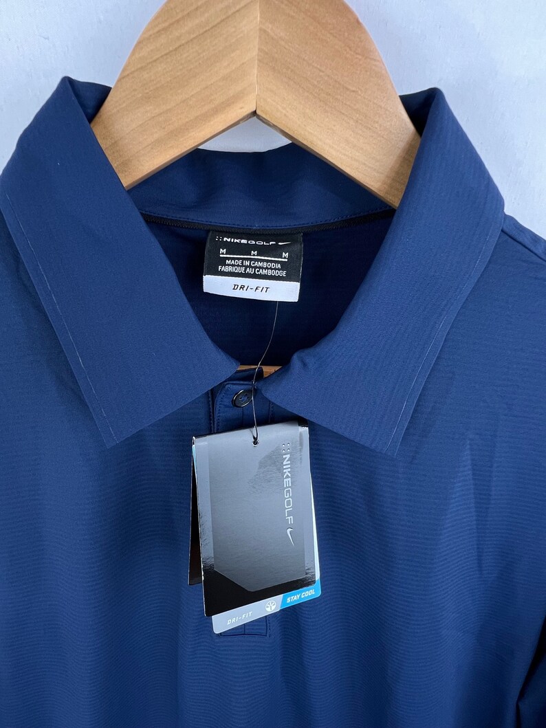 May include: A navy blue golf shirt with a white tag that says "Nikegolf" and "Dri-Fit Stay Cool". The shirt has a button-up collar and a small pocket on the left chest.
