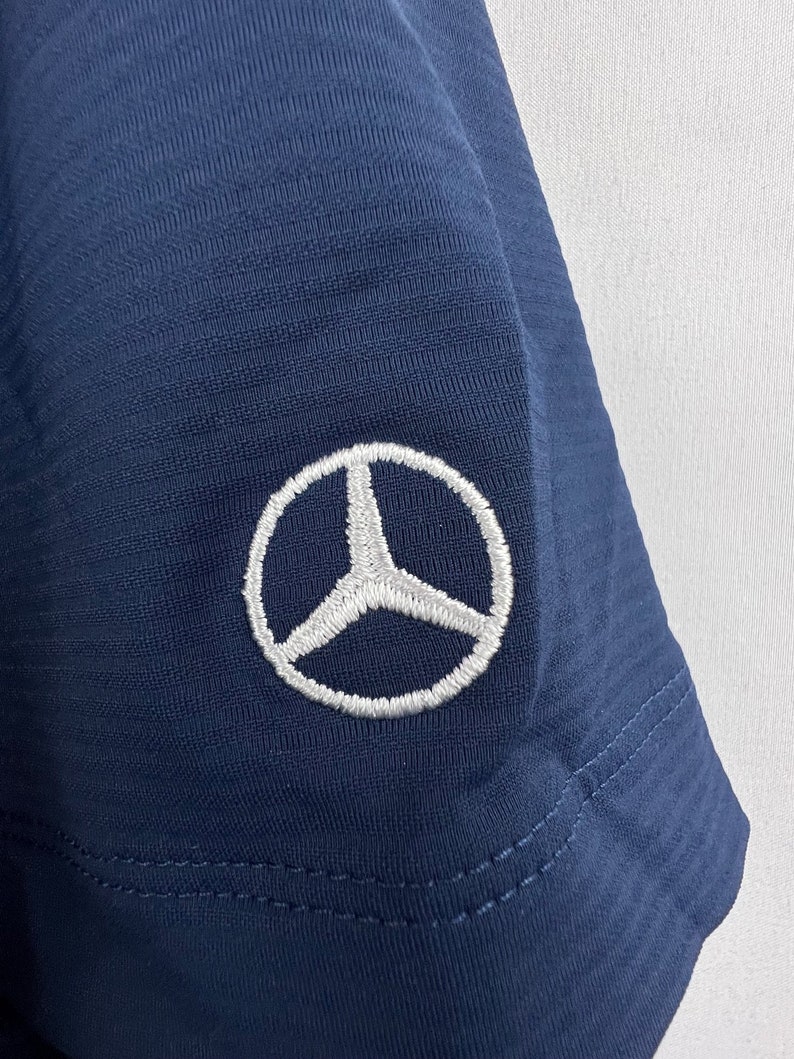 May include: A navy blue t-shirt with a white embroidered Mercedes-Benz logo on the sleeve.