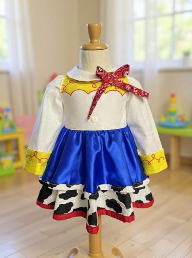 May include: A children's costume dress with a white top, blue skirt, and cow print bottom. The dress features a red bandana bow and yellow accents. The dress is displayed on a mannequin.