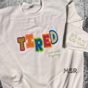 May include: White sweatshirt with the word "TIRED" in colorful block letters and the text "but still trying" in green cursive. The sweatshirt also has the text "IT'S OK TO SLOW DOWN" in green cursive on the sleeve.