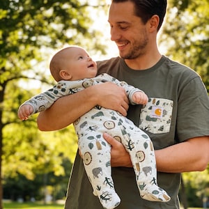 May include: A baby in a light-colored onesie with a safari animal print is held by a person wearing a green t-shirt. The onesie features lions, elephants, and other animals. The t-shirt has a pocket with a logo.