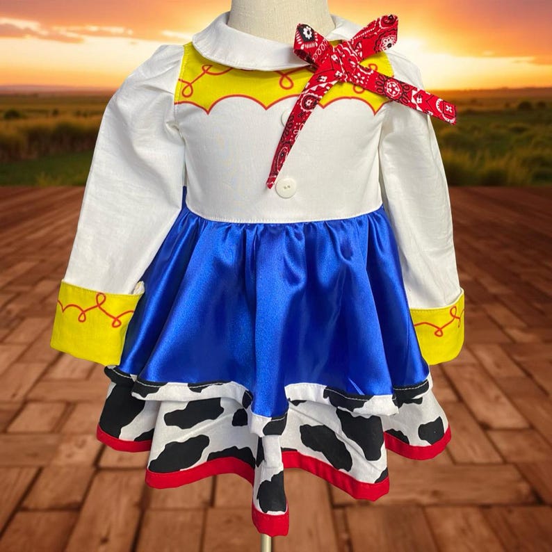 May include: A white, blue, and cow-print dress with a red bandana bow. The dress features a white collar, long sleeves, and a blue satin skirt. The bottom of the dress has a cow-print design with red trim. Yellow accents are on the sleeves.